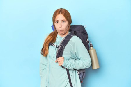 Téléchargez les photos : Voyageur rousse avec sac à dos et tapis en studio haussant les épaules et les yeux ouverts confus. - en image libre de droit