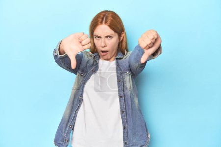 Photo for Redhead young woman on blue background showing thumb down and expressing dislike. - Royalty Free Image