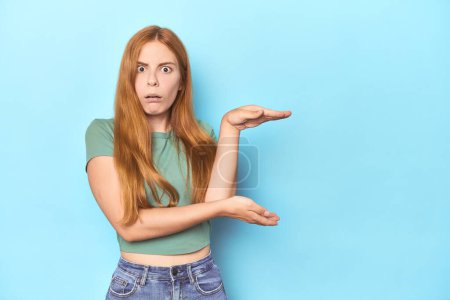 Photo for Redhead young woman on blue background shocked and amazed holding a copy space between hands. - Royalty Free Image