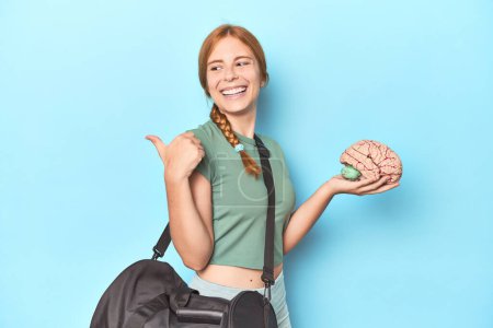 Photo for Sporty redhead with brain model on blue background points with thumb finger away, laughing and carefree. - Royalty Free Image