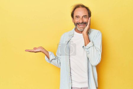 Téléchargez les photos : Homme d'âge moyen caucasien en studio jaune tient l'espace de copie sur une paume, garder la main sur la joue. Incroyable et ravi. - en image libre de droit