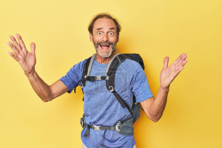 Photo for Middle-aged hiker with backpack in studio receiving a pleasant surprise, excited and raising hands. - Royalty Free Image