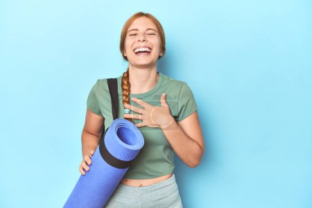 Téléchargez les photos : Rousse jeune femme tenant tapis de yoga en studio rit à haute voix en gardant la main sur la poitrine. - en image libre de droit