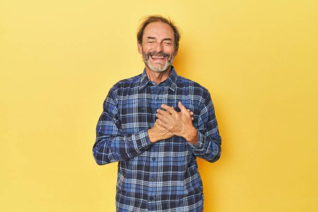 Téléchargez les photos : Homme d'âge moyen caucasien en studio jaune riant en gardant la main sur le cœur, concept de bonheur. - en image libre de droit