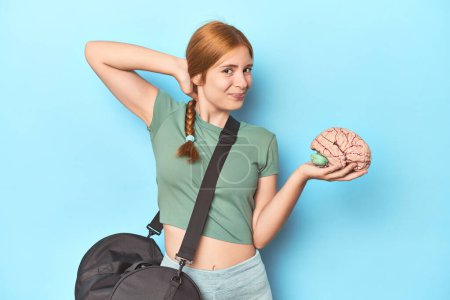 Photo for Sporty redhead with brain model on blue background touching back of head, thinking and making a choice. - Royalty Free Image