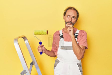 Photo for Man painting yellow backdrop in studio looking sideways with doubtful and skeptical expression. - Royalty Free Image