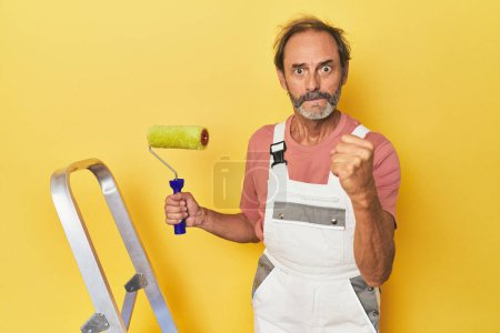 Foto de Hombre pintando fondo amarillo en estudio mostrando puño a cámara, expresión facial agresiva. - Imagen libre de derechos