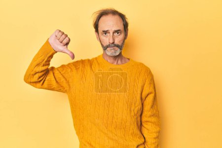 Photo for Caucasian middle-aged man in yellow studio showing a dislike gesture, thumbs down. Disagreement concept. - Royalty Free Image