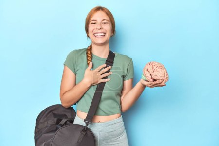 Photo for Sporty redhead with brain model on blue background laughs out loudly keeping hand on chest. - Royalty Free Image