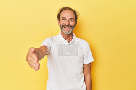 Photo for Caucasian middle-aged man in yellow studio stretching hand at camera in greeting gesture. - Royalty Free Image