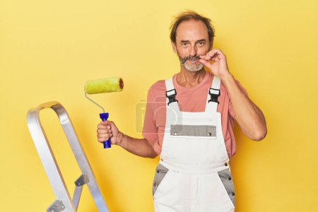 Foto de Hombre pintando fondo amarillo en estudio con los dedos en los labios manteniendo un secreto. - Imagen libre de derechos