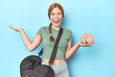 Photo for Sporty redhead with brain model on blue background receiving a pleasant surprise, excited and raising hands. - Royalty Free Image