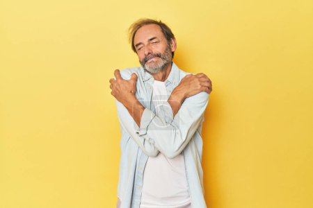 Photo for Caucasian middle-aged man in yellow studio hugs, smiling carefree and happy. - Royalty Free Image