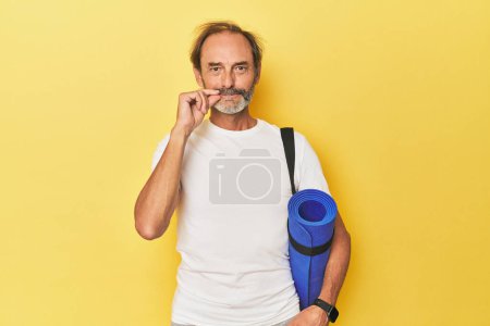 Foto de Hombre con esterilla de yoga en estudio amarillo con los dedos en los labios manteniendo un secreto. - Imagen libre de derechos