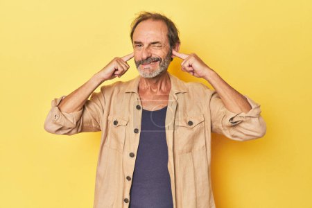 Photo for Caucasian middle-aged man in yellow studio covering ears with fingers, stressed and desperate by a loudly ambient. - Royalty Free Image