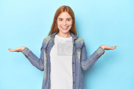 Téléchargez les photos : Rousse jeune femme sur fond bleu fait échelle avec les bras, se sent heureux et confiant. - en image libre de droit