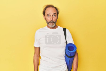 Photo for Man with yoga mat in yellow studio shrugs shoulders and open eyes confused. - Royalty Free Image
