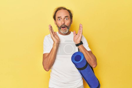 Téléchargez les photos : Homme avec tapis de yoga en studio jaune surpris et choqué. - en image libre de droit