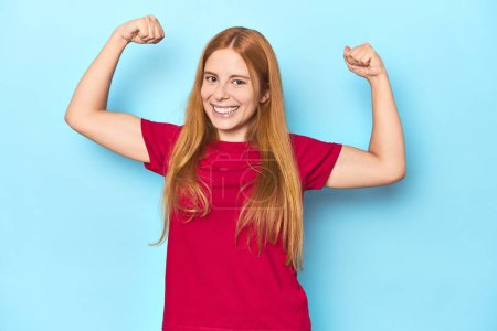 Téléchargez les photos : Jeune femme rousse sur fond bleu montrant un geste de force avec des bras, symbole du pouvoir féminin - en image libre de droit