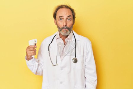 Foto de Doctor sosteniendo termómetro infrarrojo en estudio encoge hombros y ojos abiertos confundido. - Imagen libre de derechos