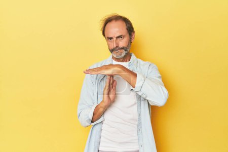 Foto de Caucásico hombre de mediana edad en estudio amarillo mostrando un gesto de tiempo de espera. - Imagen libre de derechos