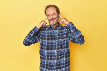 Téléchargez les photos : Homme d'âge moyen caucasien dans un studio jaune couvrant les oreilles avec les doigts, stressé et désespéré par une ambiance bruyante. - en image libre de droit