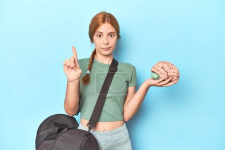 Photo for Sporty redhead with brain model on blue background showing number one with finger. - Royalty Free Image