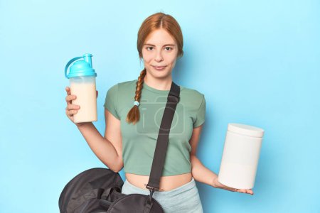 Photo for Athletic redhead holding protein shake and bottle in blue studio - Royalty Free Image