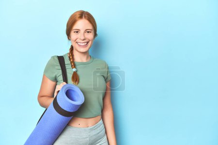 Photo for Sporty redhead woman carrying yoga mat in blue studio background - Royalty Free Image
