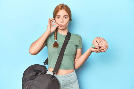 Photo for Sporty redhead with brain model on blue background with fingers on lips keeping a secret. - Royalty Free Image