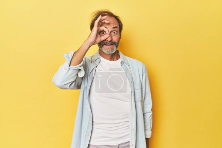 Foto de Caucásico de mediana edad hombre en amarillo estudio emocionado mantener el gesto bien en el ojo. - Imagen libre de derechos