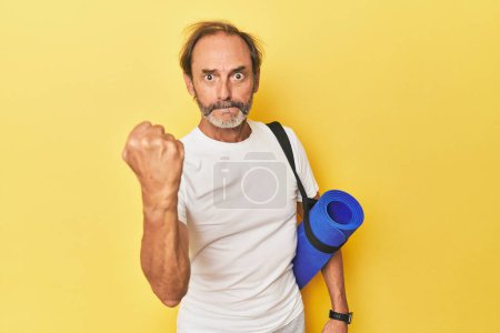 Téléchargez les photos : Homme avec tapis de yoga en studio jaune montrant poing à caméra, expression faciale agressive. - en image libre de droit