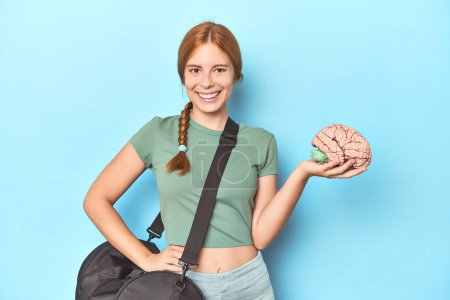 Photo for Sporty redhead with brain model on blue background happy, smiling and cheerful. - Royalty Free Image