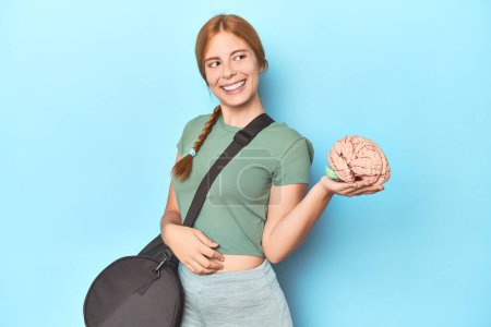 Photo for Sporty redhead with brain model on blue background looks aside smiling, cheerful and pleasant. - Royalty Free Image