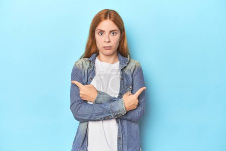 Photo for Redhead young woman on blue background points sideways, is trying to choose between two options. - Royalty Free Image