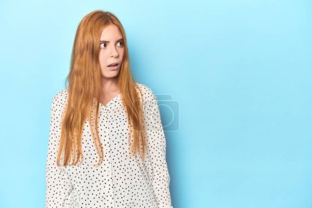 Photo for Redhead young woman on blue background being shocked because of something she has seen. - Royalty Free Image