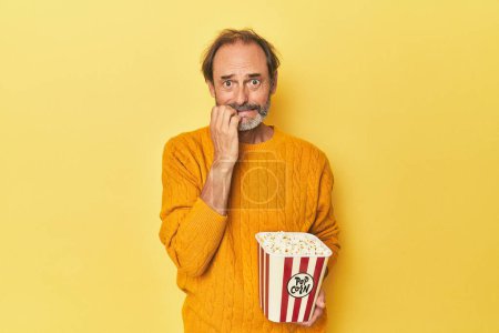 Téléchargez les photos : Homme appréciant le pop-corn en studio jaune mordant les ongles, nerveux et très anxieux. - en image libre de droit