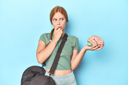 Photo for Sporty redhead with brain model on blue background relaxed thinking about something looking at a copy space. - Royalty Free Image