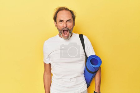 Foto de Hombre con esterilla de yoga en estudio amarillo gritando muy enojado y agresivo. - Imagen libre de derechos
