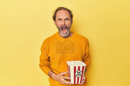 Photo for Man enjoying popcorn in yellow studio screaming very angry and aggressive. - Royalty Free Image