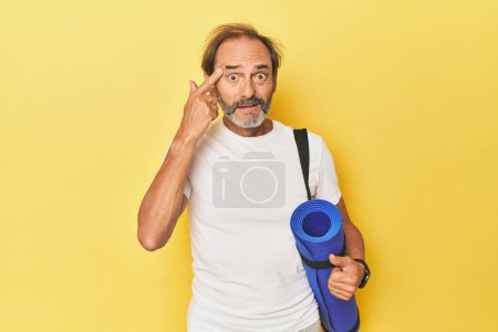Foto de Hombre con esterilla de yoga en estudio amarillo mostrando un gesto decepcionante con dedo índice. - Imagen libre de derechos