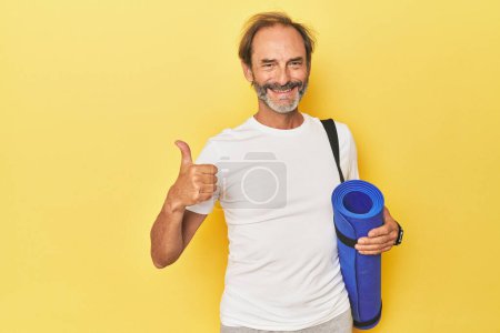 Téléchargez les photos : Homme avec tapis de yoga en studio jaune souriant et levant le pouce - en image libre de droit