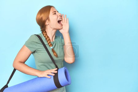 Téléchargez les photos : Rousse jeune femme tenant tapis de yoga en studio criant et tenant la paume près de la bouche ouverte. - en image libre de droit
