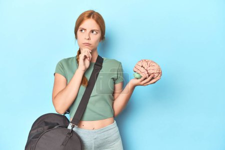 Photo for Sporty redhead with brain model on blue background looking sideways with doubtful and skeptical expression. - Royalty Free Image
