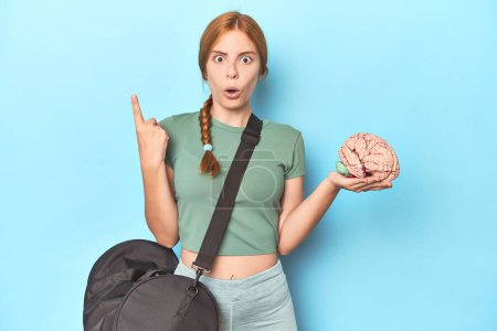 Photo for Sporty redhead with brain model on blue background having some great idea, concept of creativity. - Royalty Free Image