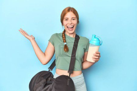 Photo for Redhead holding protein shake in blue studio receiving a pleasant surprise, excited and raising hands. - Royalty Free Image