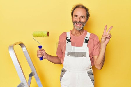 Téléchargez les photos : Homme peignant fond jaune en studio joyeux et insouciant montrant un symbole de paix avec les doigts. - en image libre de droit