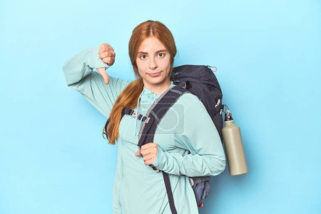 Photo for Redhead traveler with backpack and mat in studio showing a dislike gesture, thumbs down. Disagreement concept. - Royalty Free Image