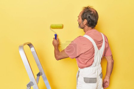 Téléchargez les photos : Homme d'âge moyen peint un mur jaune avec échelle - en image libre de droit
