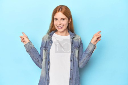 Photo for Redhead young woman on blue background pointing to different copy spaces, choosing one of them, showing with finger. - Royalty Free Image